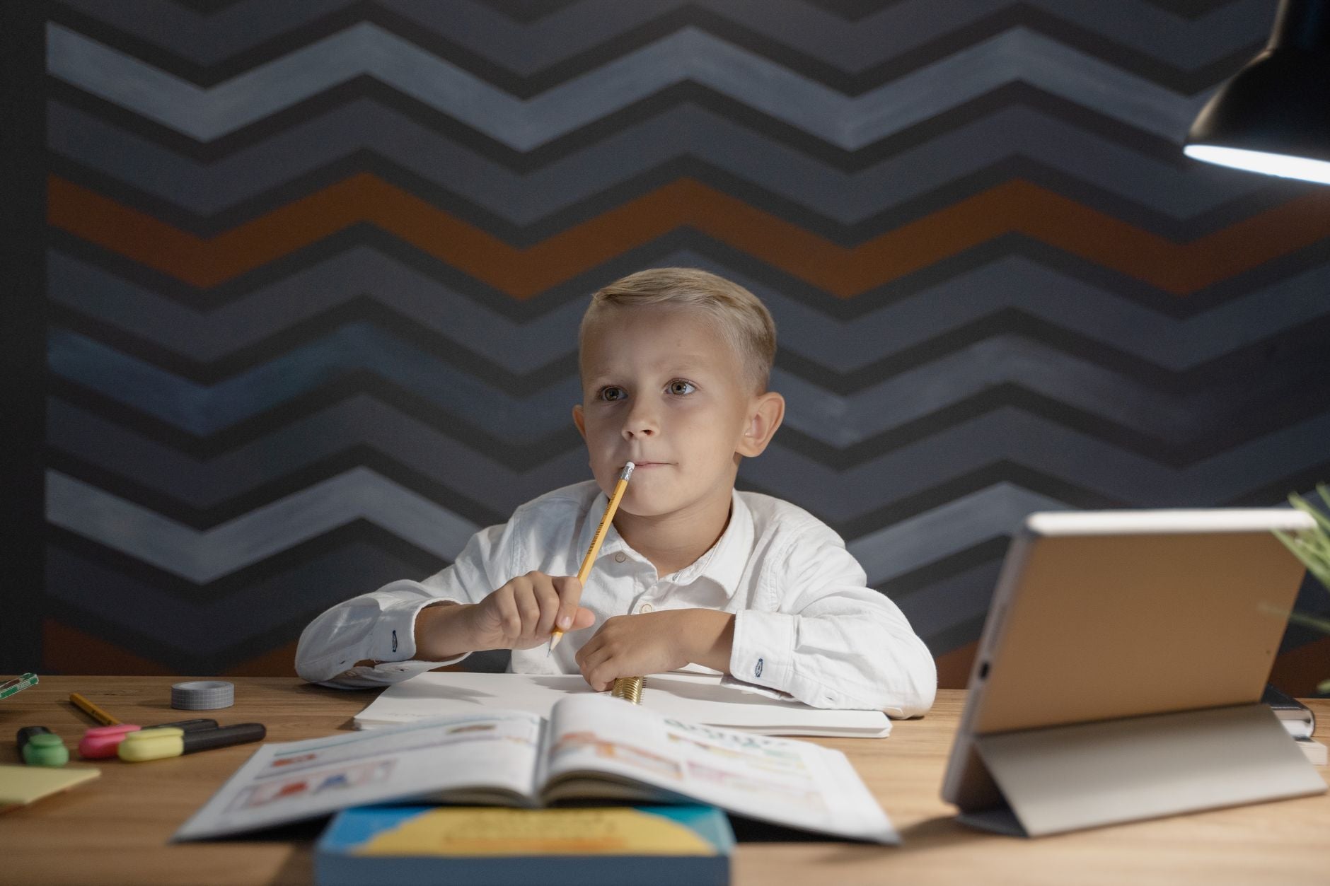 Young boy sitting at desk with open books and tablet, pondering while doing homework under a desk lamp.