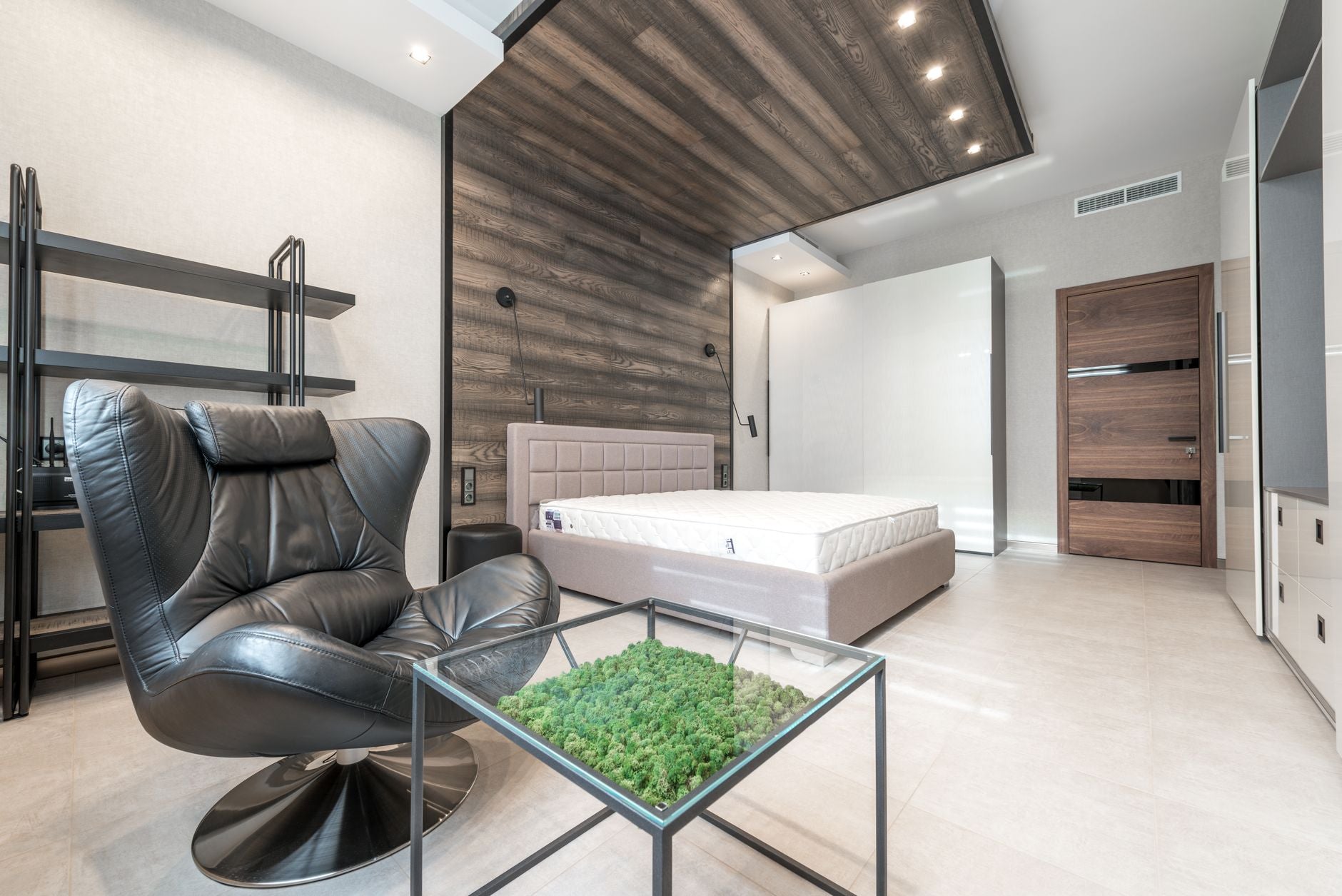 Leather armchair placed near table and bed with mattress in spacious bedroom with door and white closet in modern apartment