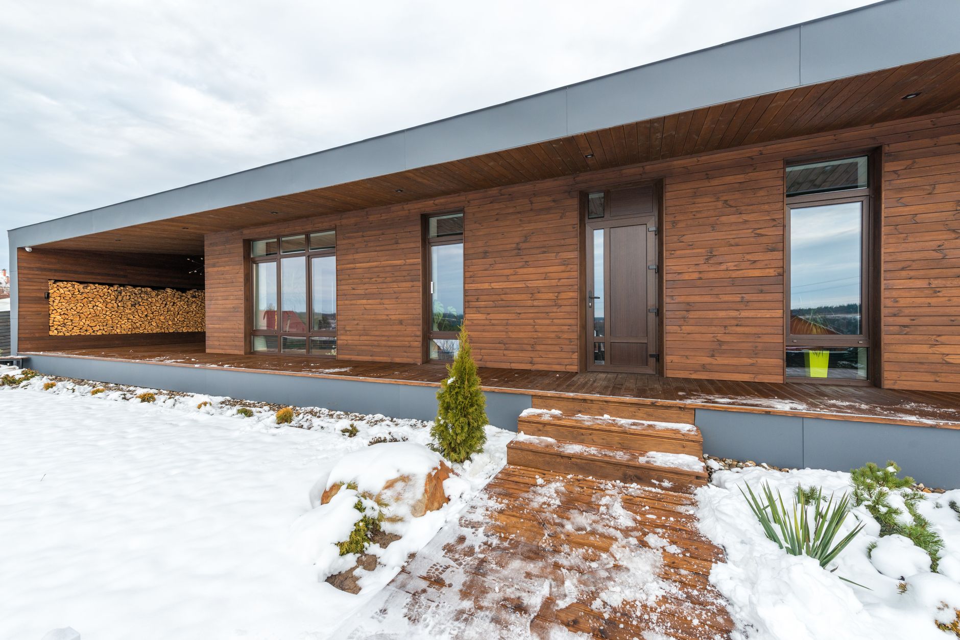 Contemporary wooden house with snow-covered yard and stacked wood for winter.