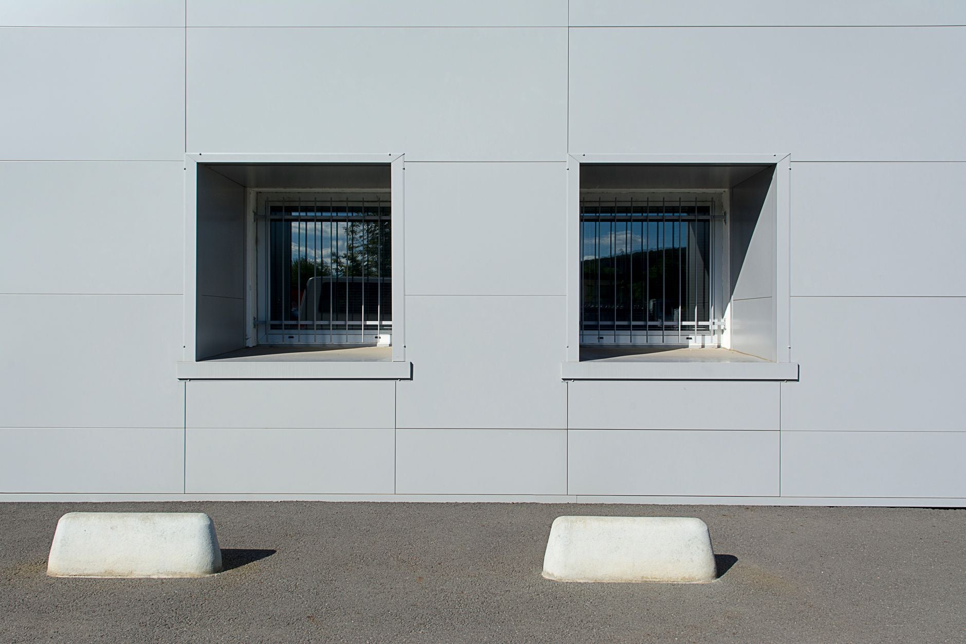 Minimalist urban facade with two sunlit windows and a concrete barrier in front.