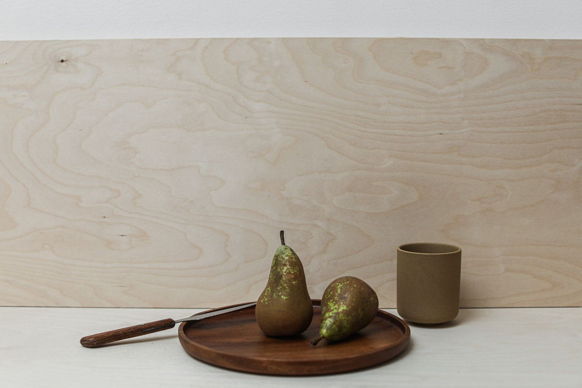 A minimalist still life featuring two pears on a wooden tray with a cup, set against a neutral wood background.