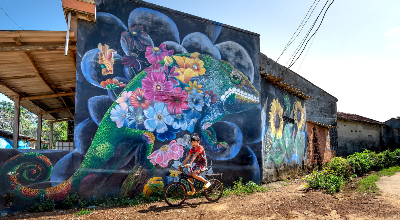 Vibrant street mural featuring a floral iguana with a child cycling past.